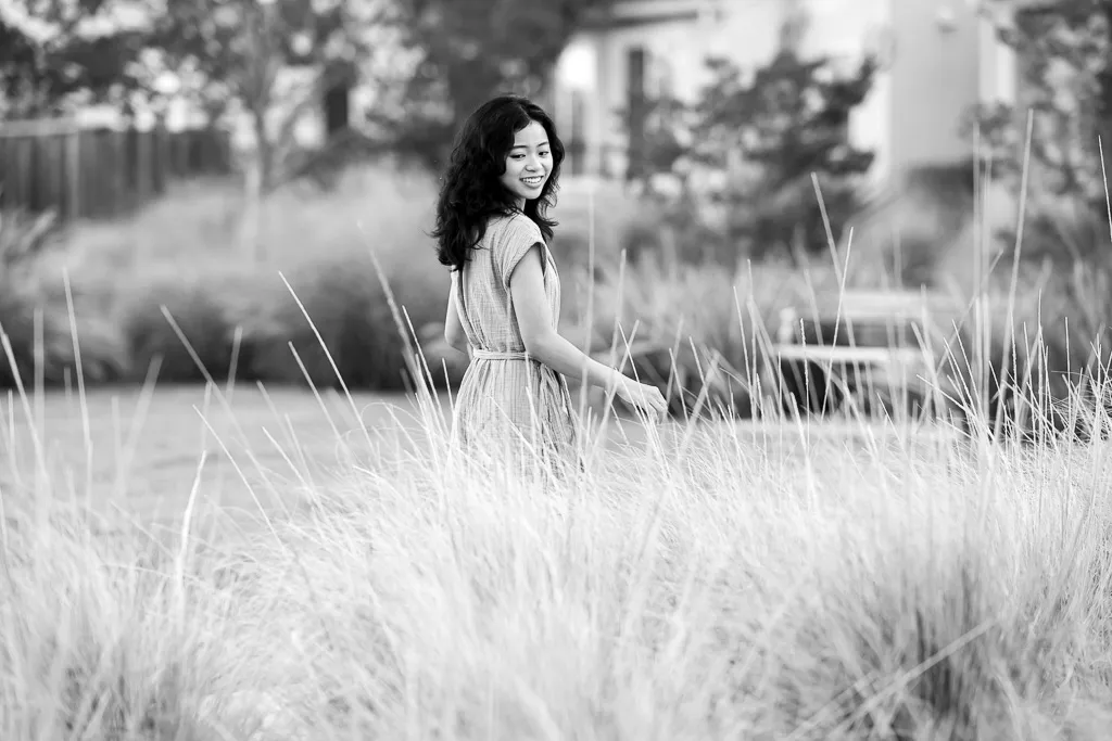 Black and white portrait of a young woman standing in tall grass, looking back over her shoulder